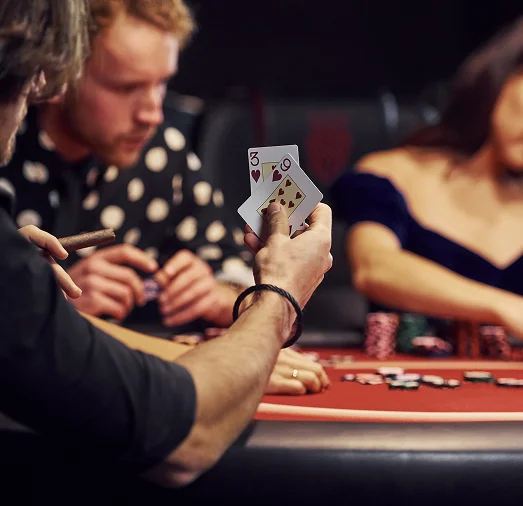 Drie mensen rond een pokertafel, een man met kaarten in zijn hand en een sigaar in zijn mond. Een andere man en een vrouw kijken geconcentreerd naar hun kaarten